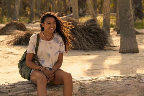 Madison Bailey como Kiara " Kie" en Outer Banks sentada en un tronco en la playa mirando hacia la derecha mientras el viento sopla en su cabello