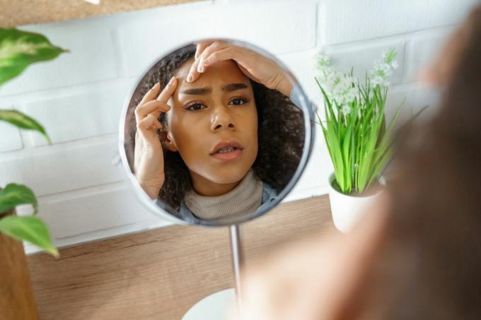 afrikaans tienermeisje maakt zich zorgen over het probleem van de gezichtshuid kijk in de spiegel jonge vrouw aanraking gezicht squish pop zit puistje op het voorhoofd gefrustreerd door mee-eters zwart huidverzorgingsprobleem, zelfzorgconcept